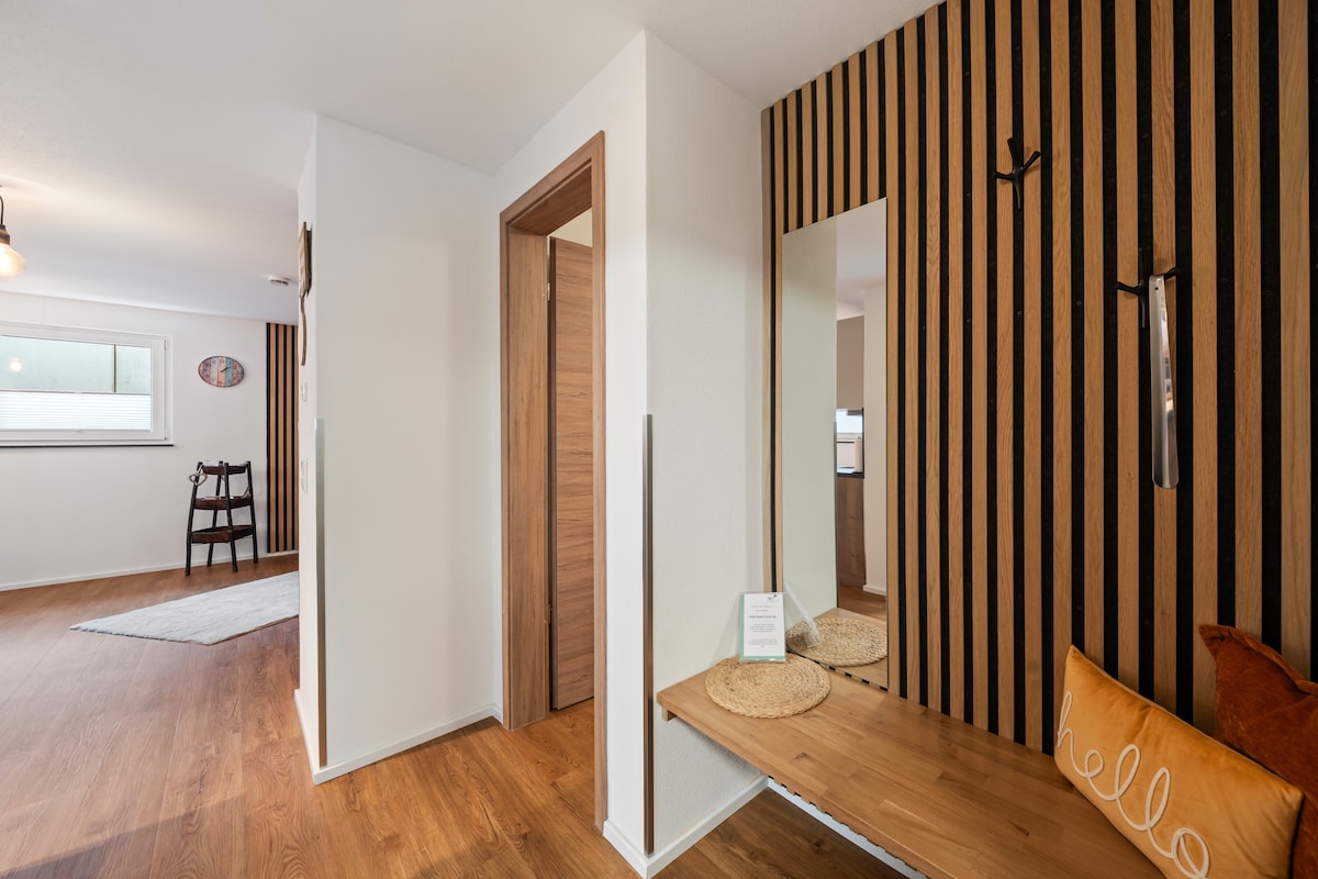 An entrance area features sleek wooden accents and a large mirror reflecting the open living space. A wooden bench is placed against the wall, accompanied by a decorative cushion. Warm lighting adds to the inviting ambiance, with a stylish rug positioned on the floor.