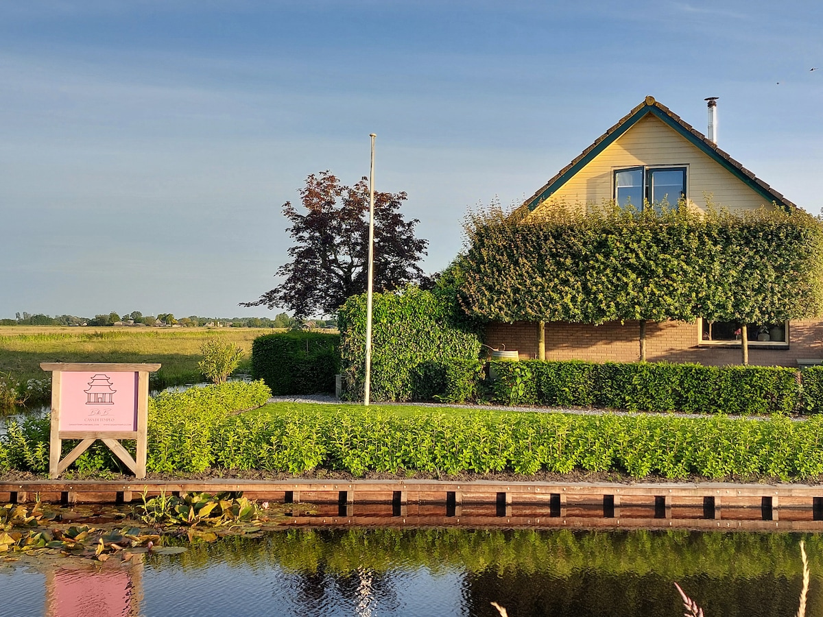 A charming house sits beside a serene waterway, surrounded by lush greenery. The structure features a sloped roof and large windows. A wooden sign with the word 'Lily' stands near the water, enhancing the tranquil rural setting.