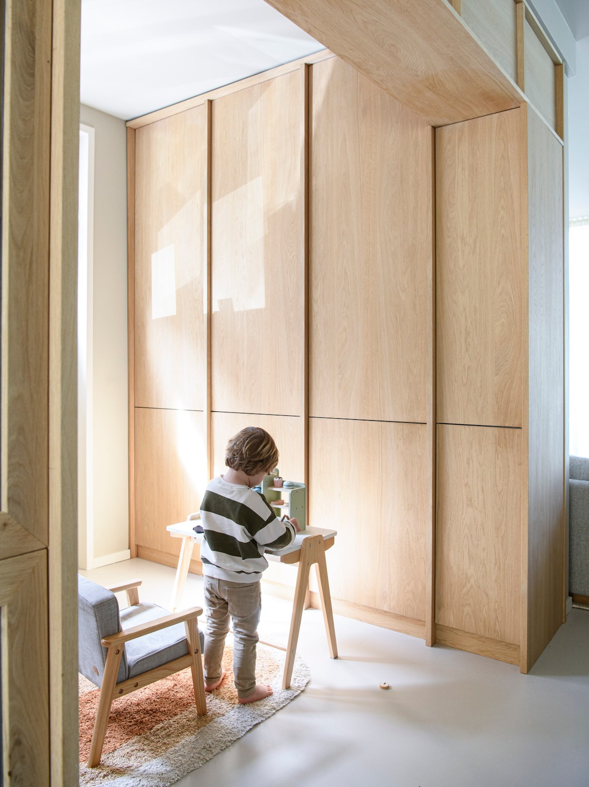 A well-lit corner shows a young child engaged in play at a small table, surrounded by warm-toned cabinetry. A comfortable chair and a soft rug add to the cozy atmosphere. Natural light filters through a nearby window, enhancing the inviting environment.