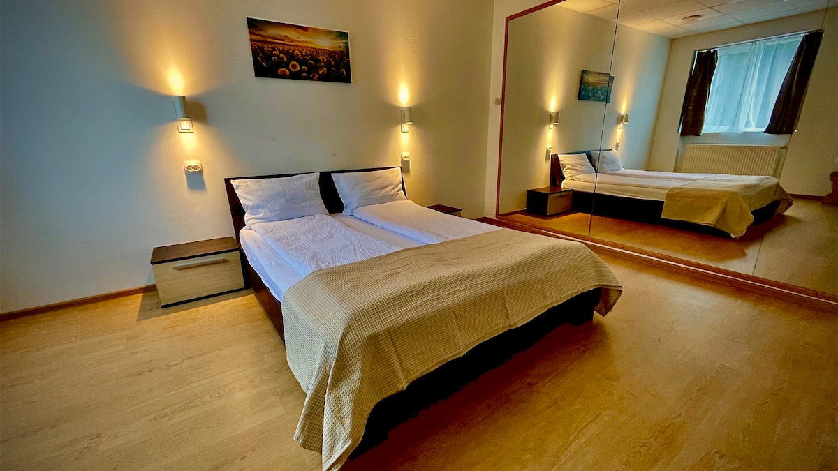 A bright bedroom features a spacious bed with neatly arranged white linens. Soft lighting from wall sconces creates a calming ambiance. A large mirror on one wall reflects the room, enhancing the sense of space. A framed nature photograph hangs above a small bedside table.