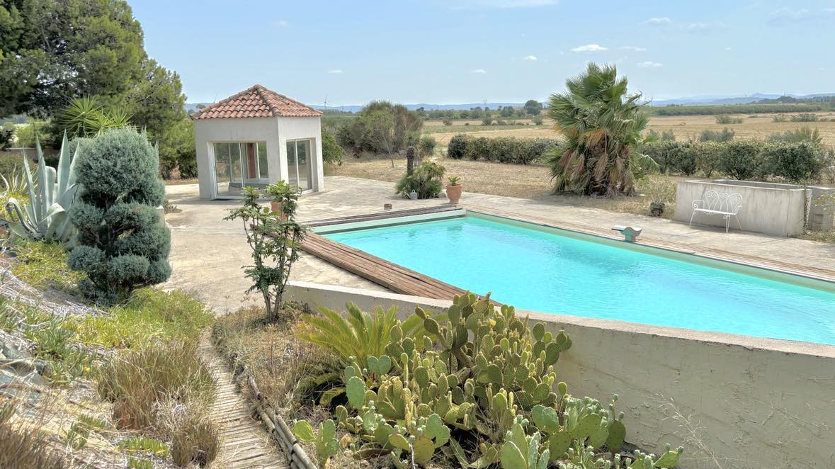 A spacious outdoor area features a turquoise swimming pool surrounded by a stone terrace. Lush greenery and palm trees are visible in the background, creating a serene environment. A small gazebo is positioned nearby, adding to the charm of the space.