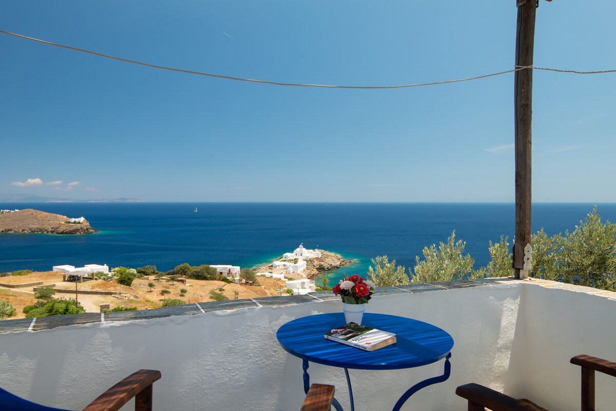 A balcony offers a serene view of the sea and nearby coastline. A small round table with blue accents is set, accompanied by wooden chairs. A flower vase and a stack of magazines are placed on the table, with clear skies and calm water creating a peaceful backdrop.