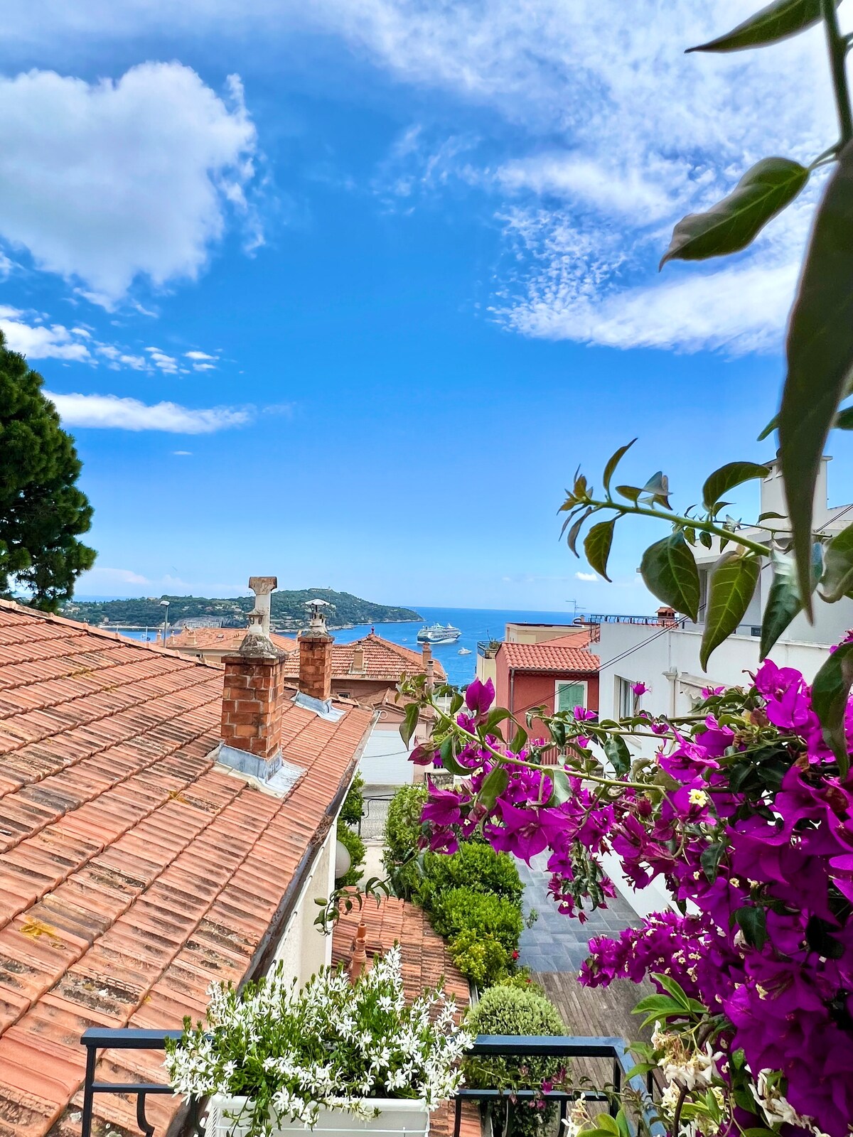 A view of terracotta rooftops is framed by vibrant bougainvillea blooms. Beyond the rooftops, the serene sea can be seen with distant hills and a bridge, all set against a clear blue sky.