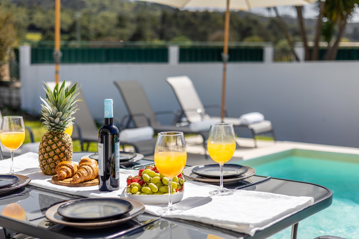A dining table is set near a pool, featuring plates with fresh fruits and croissants. Handled glasses with orange drinks stand on the table, accompanied by a bottle of wine. Sun loungers are arranged in the background, with a bright umbrella providing shade.