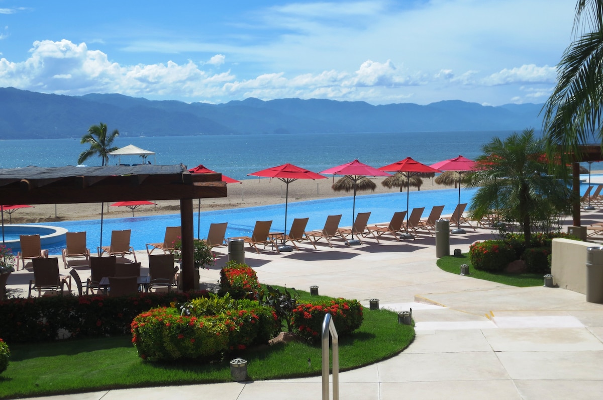 A scenic view features a sprawling beachside area with sun loungers arranged around a shimmering pool. Vibrant red umbrellas provide shade, while lush greenery and flowerbeds enhance the outdoor space. The ocean stretches in the background, framed by distant mountains under a bright blue sky.