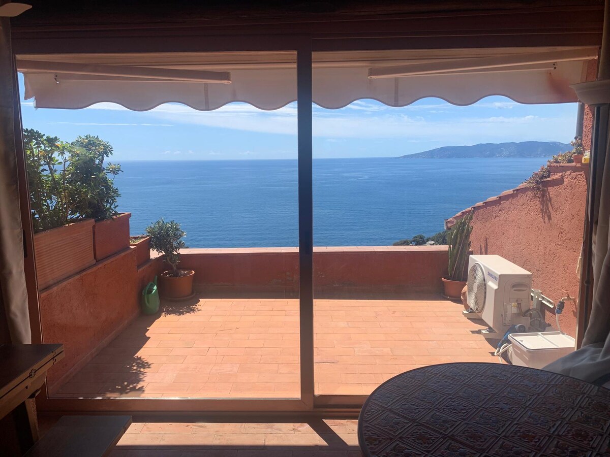 A spacious terrace is visible through glass sliding doors, featuring terracotta tile flooring and potted plants. The panoramic sea view includes the distant outline of Giglio Island under a clear blue sky. A wall-mounted air conditioning unit is positioned to the right.