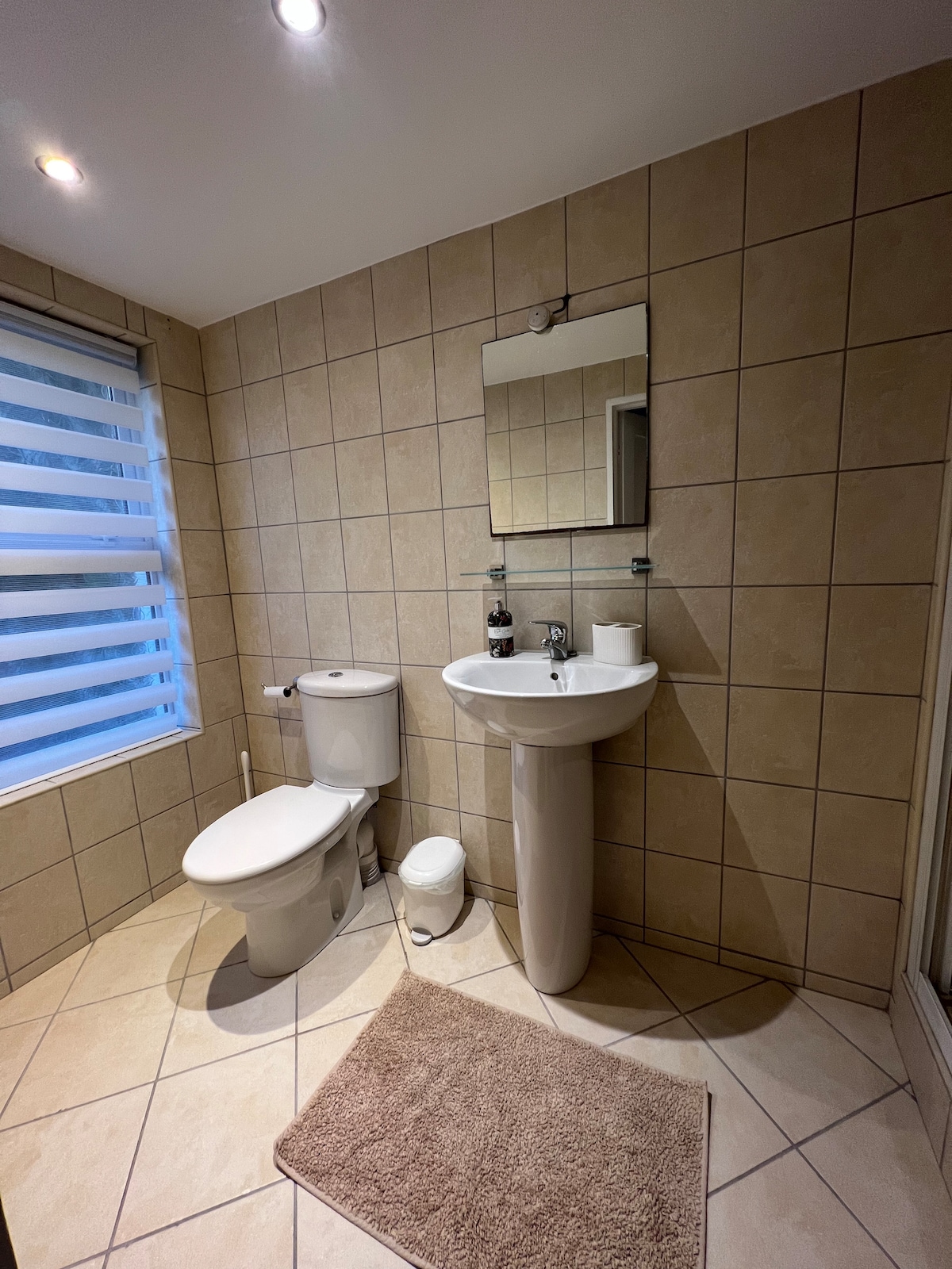A modern bathroom is presented with beige tiled walls and floor. A white pedestal sink and a matching toilet are visible. A full-length mirror hangs above the sink. Natural light enters through a window adorned with horizontal blinds, complementing the soft tones of the space.