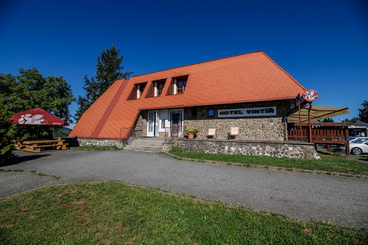 The hotel exterior features a unique triangular roof with bright orange tiles. A stone façade is complemented by a welcoming entrance and outdoor seating areas. A picnic table under a shaded umbrella is visible, surrounded by greenery, providing a relaxing spot for guests.