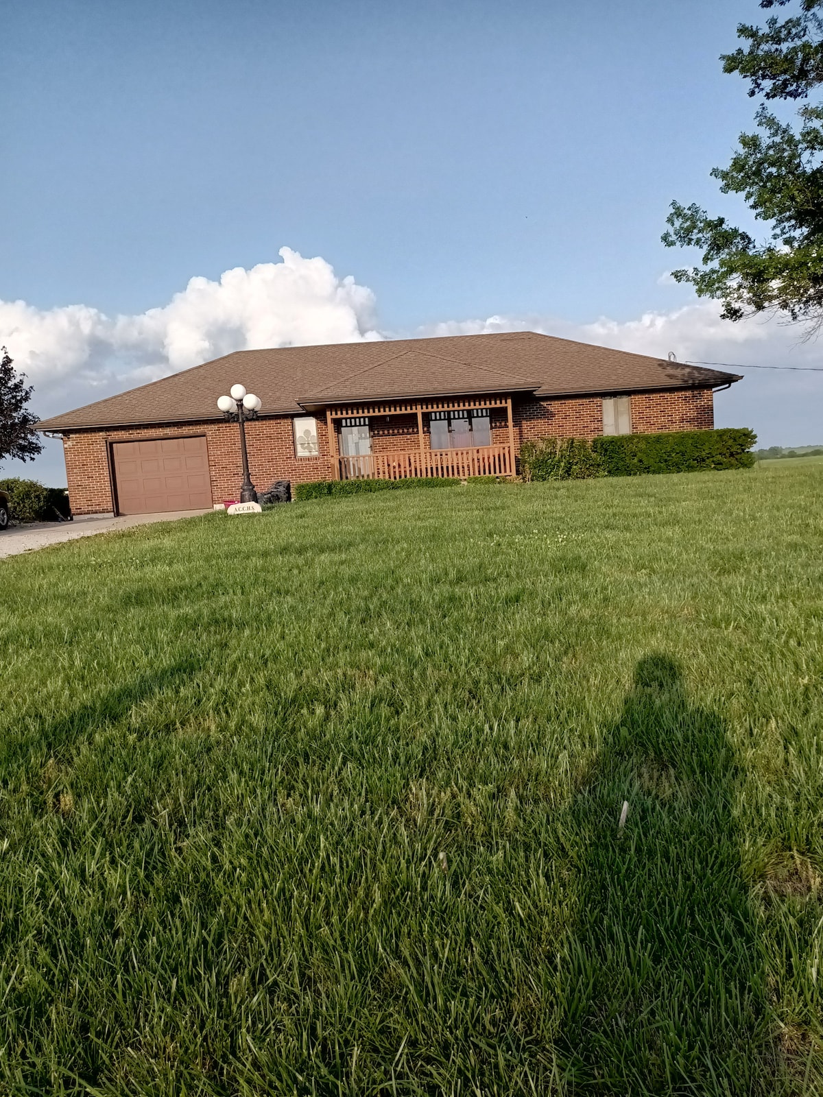 A single-story brick house features a front porch with decorative railing. Large windows allow natural light into the interior, while a well-maintained lawn with tall green grass surrounds the home. A light fixture stands near the entrance, providing illumination.