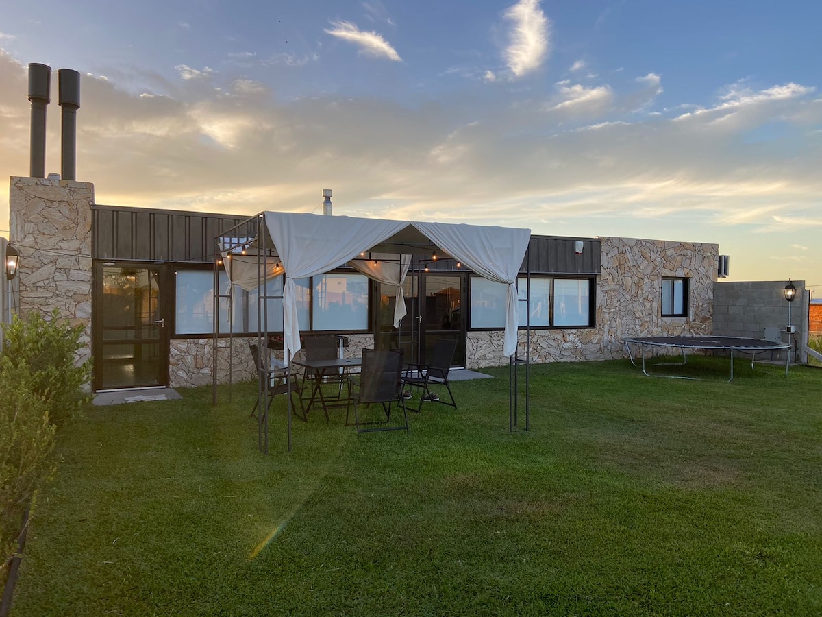 An outdoor area features a spacious lawn with green grass and a stone house. A covered seating area is equipped with dark outdoor furniture, including a table and chairs. A trampoline is positioned nearby, and soft clouds can be seen in the clear sky.