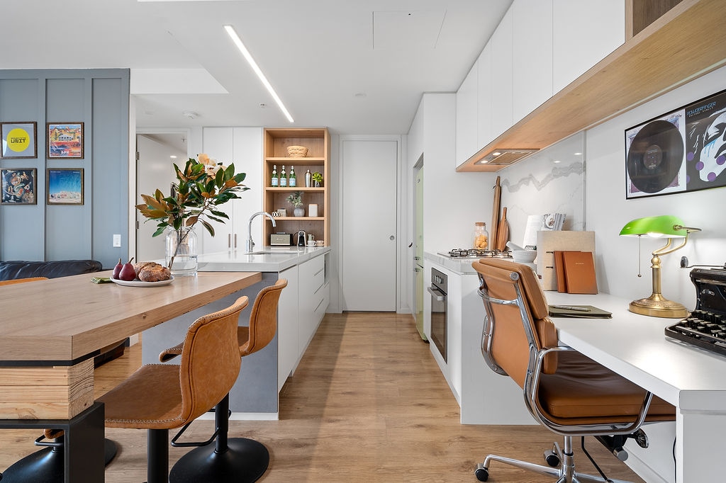 A modern kitchen features sleek cabinetry and a minimalistic design, complemented by a wooden peninsula with high stools. A desk area with a vintage typewriter and a green desk lamp is visible. Natural light is illuminated through a nearby window.