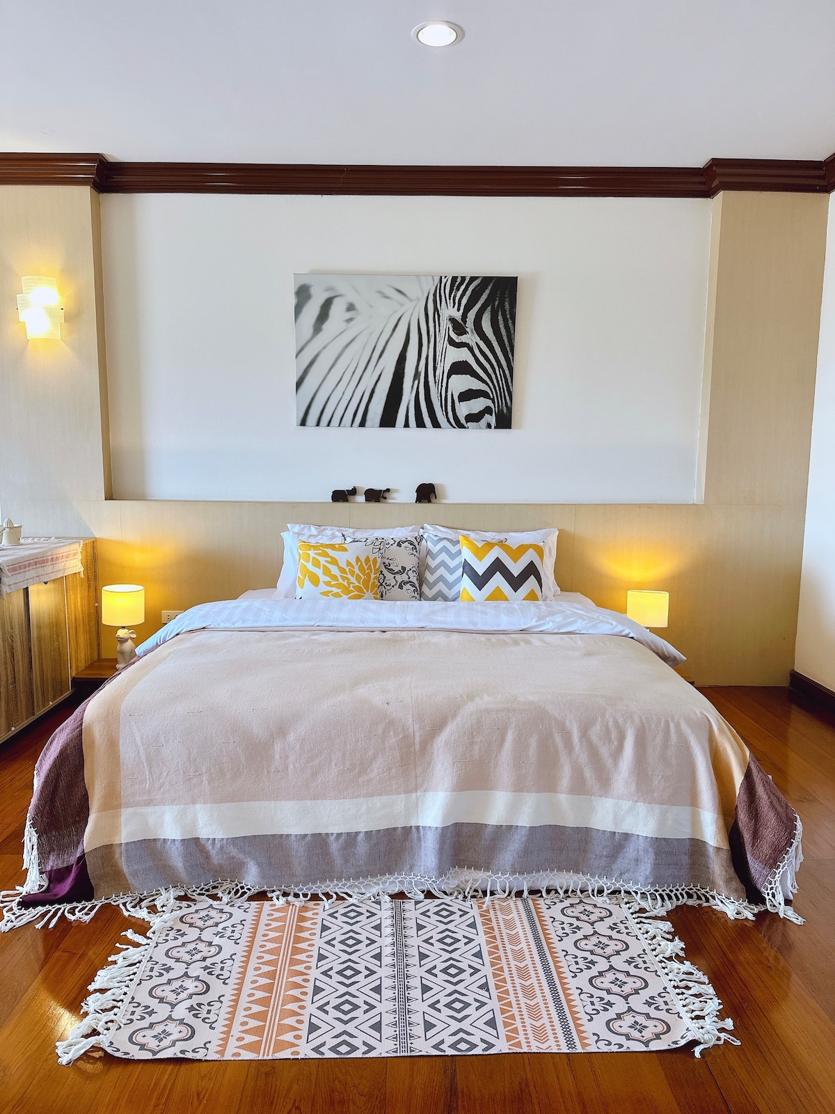A spacious sleeping area features a soft bed adorned with layered blankets, complemented by colorful cushions and a decorative rug. The walls are accented by a zebra-print artwork, while gentle lighting from bedside lamps enhances the room's inviting atmosphere.