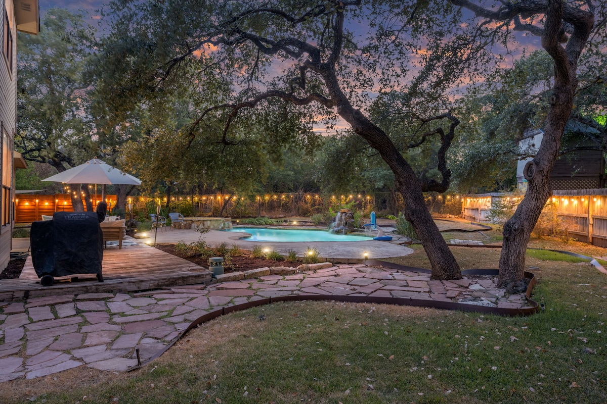 A serene backyard scene is illuminated by soft string lights, highlighting the heated pool surrounded by lush greenery. Comfortable seating areas are positioned for relaxation, while a patio umbrella offers shade. The winding stone path invites guests to explore the inviting outdoor oasis.
