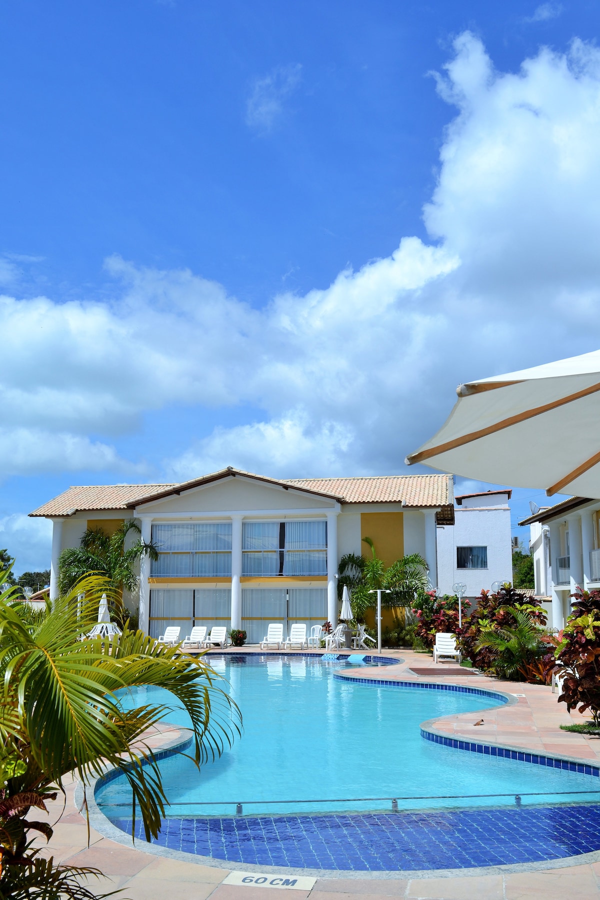 A serene outdoor area features a clear swimming pool surrounded by lush greenery and sun loungers. The two-story building in the background showcases large windows and a welcoming design under a bright blue sky, enhancing the tranquil atmosphere.