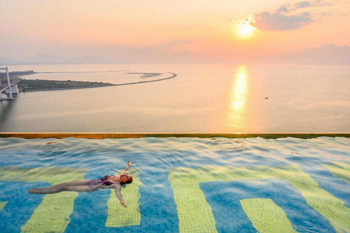 An infinity pool is positioned on a rooftop, offering breathtaking views of the ocean and mountains at sunset. The calm water reflects the warm colors of the sky, while a swimmer enjoys the serene atmosphere surrounded by distant landscapes.