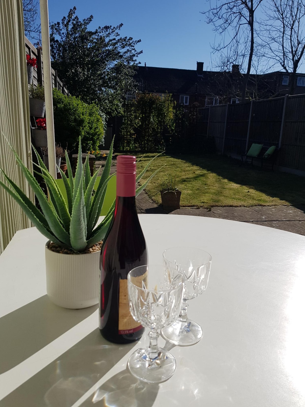 A table is set outside with a bottle of red wine and two crystal glasses, emphasizing a relaxing outdoor experience. In the background, the green lawn of a private garden is visible, surrounded by a high fence, under a clear blue sky.