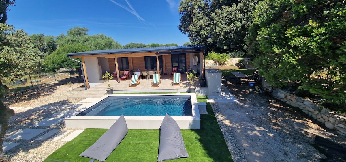 The outdoor area features a serene swimming pool surrounded by a well-maintained lawn. Two gray lounge cushions are positioned on the grass, and several wooden chairs are visible near the pool. The house, with a wooden facade, is framed by lush greenery and clear blue skies.