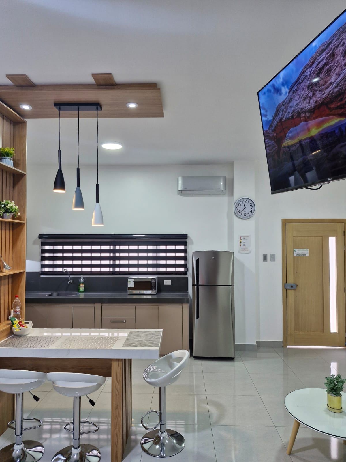A modern kitchen space features sleek cabinetry and a stone countertop. Three bar stools are positioned at the kitchen island. A large refrigerator and a wall-mounted television are visible, along with natural light filtering through window treatments.