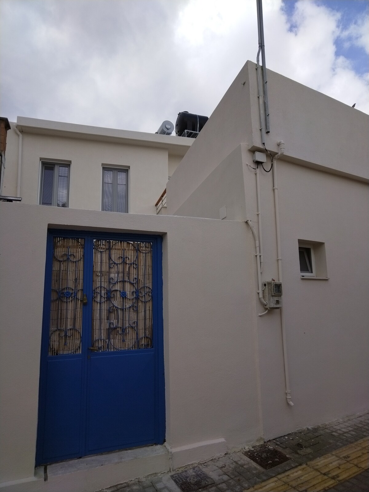 A charming exterior of a house is presented, featuring a bright blue door detailed with ornate ironwork. The clean white walls and visible windows create a simple yet inviting entrance. A cobblestone pathway leads up to the door, surrounded by a light, tranquil atmosphere.