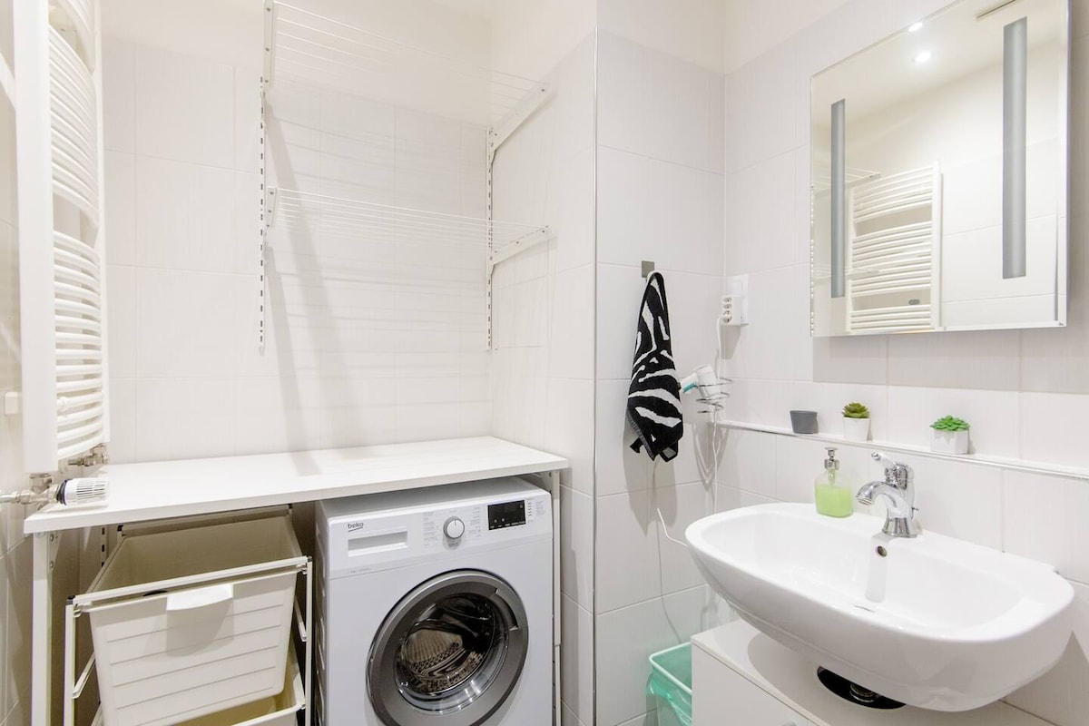 A modern bathroom is fitted with a washer and a white countertop. A circular basin is positioned on the countertop, accompanied by a mirror and minimalistic decor. A drying rack and a small storage cabinet are mounted on the walls, creating an organized and functional space.