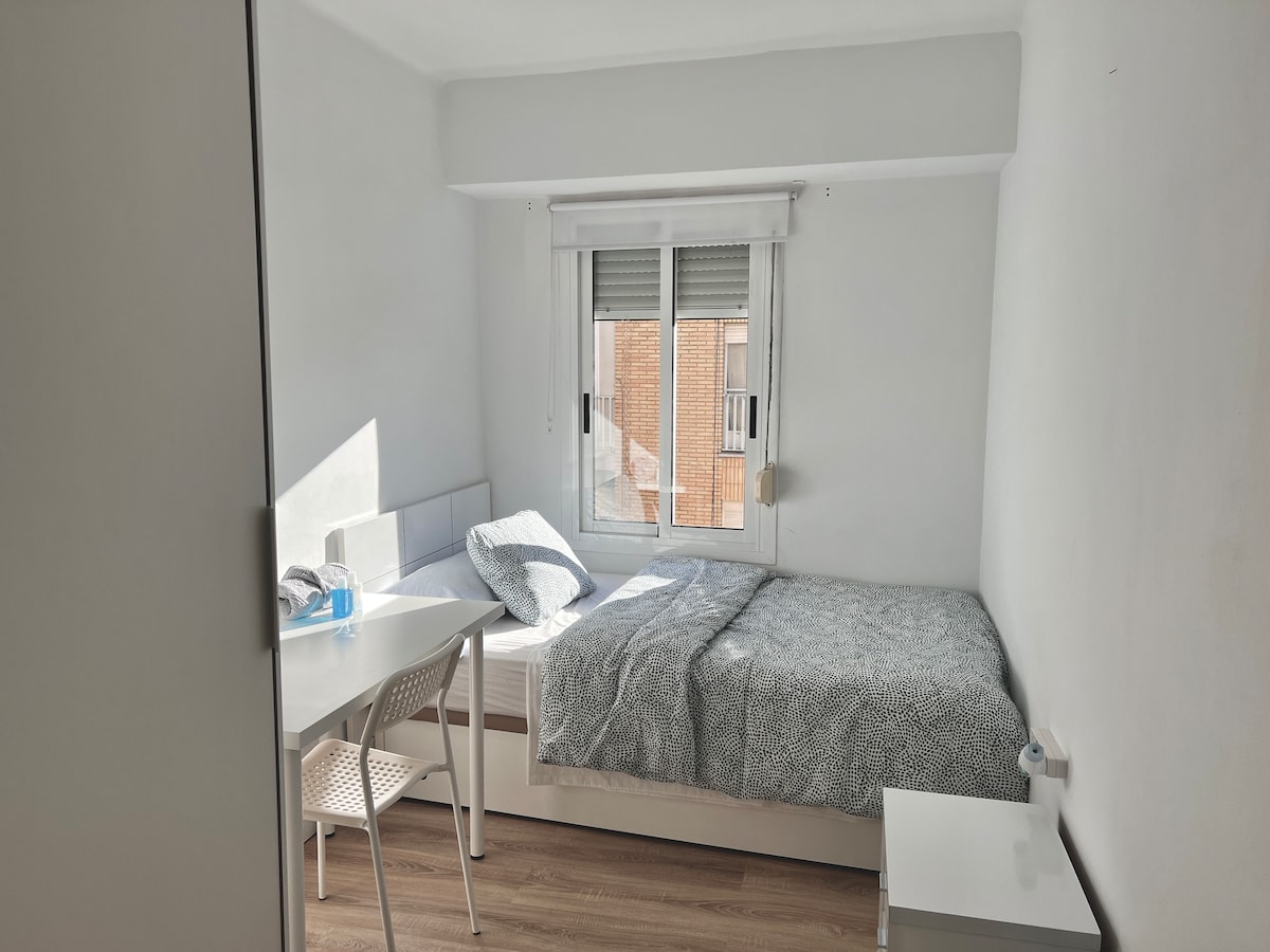 A bright and clean bedroom features a spacious bed with a grey patterned blanket. A desk and chair provide a study area, and a window offers natural light. A small nightstand is positioned beside the bed, enhancing the room's functionality.