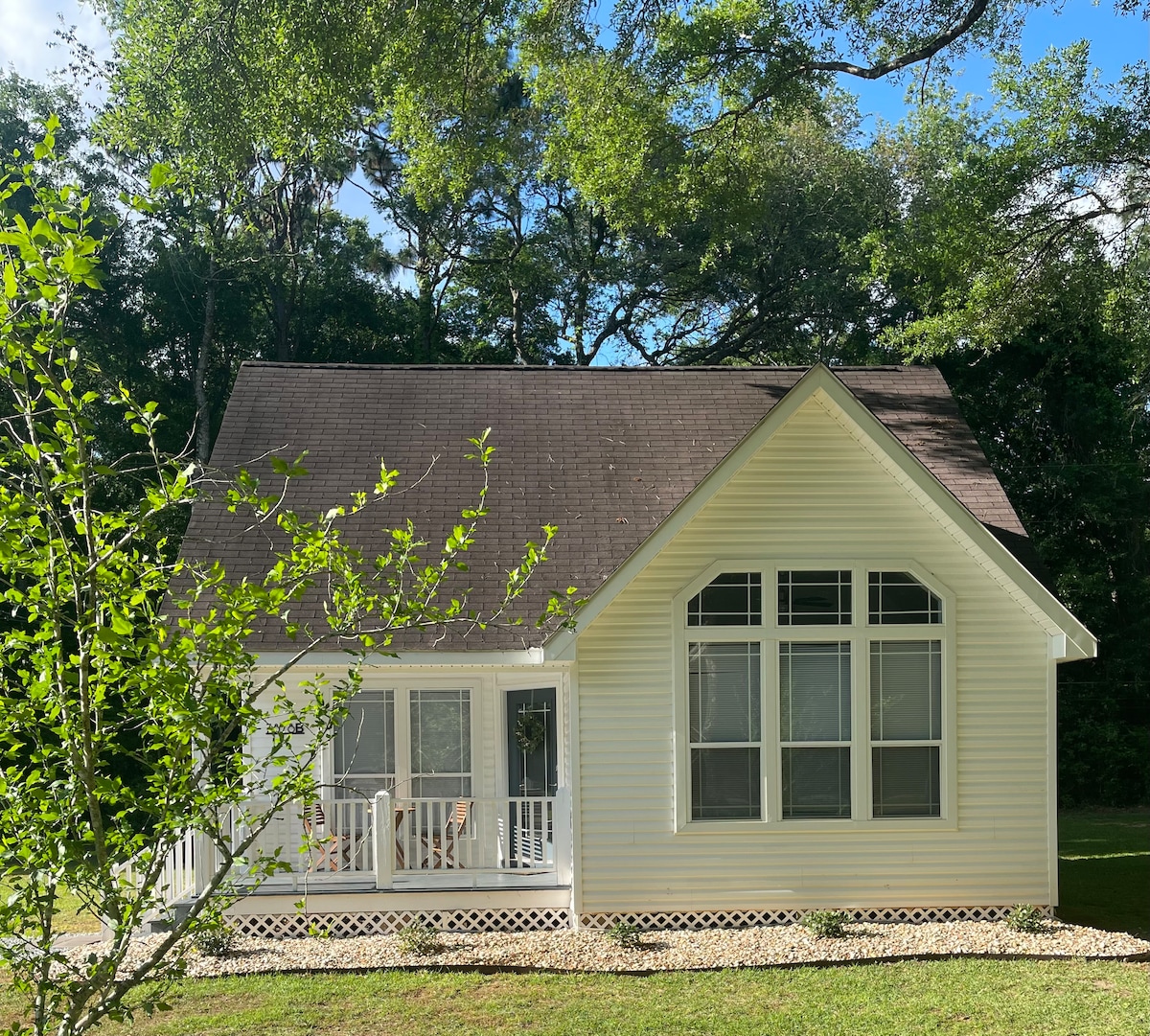 A cottage exterior is visible, featuring a light-colored façade and large windows that allow natural light inside. A front porch is present, with steps leading up from a stone and gravel area. Surrounding greenery includes oak trees, creating a natural and serene setting.