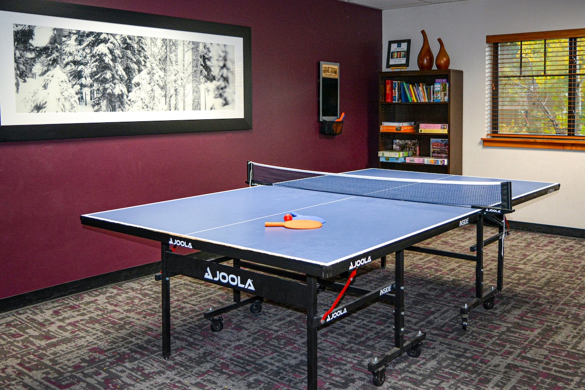 A ping pong table is positioned in a game room with a burgundy accent wall. Nearby shelves hold books and a decorative arrangement, while a large framed black-and-white photograph adorns another wall. Natural light filters through a window with blinds, creating a bright atmosphere.
