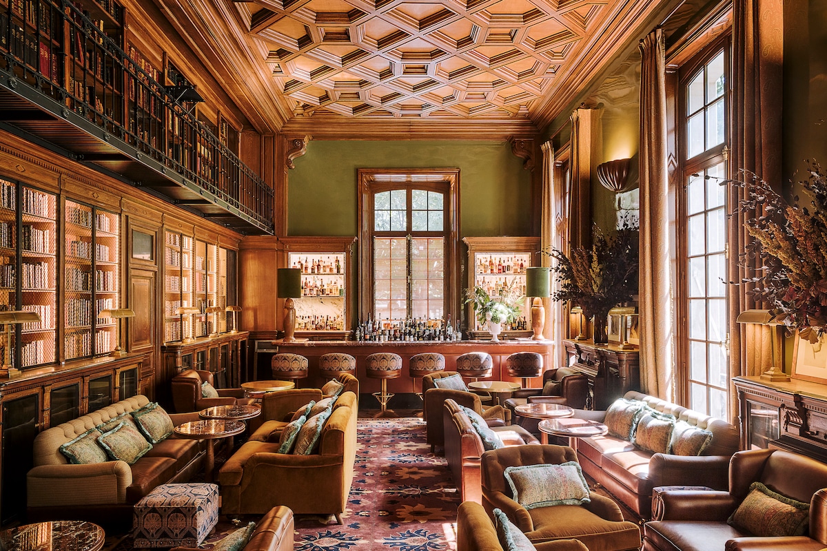 A spacious lounge area is characterized by wood-paneled walls and a coffered ceiling. Comfortable seating, including plush armchairs and sofas, is arranged around low tables. Large windows allow natural light to fill the room, while shelves of books add a touch of sophistication.