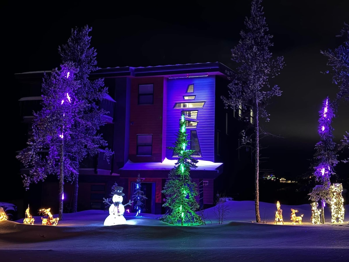 A three-story home is showcased under nighttime lighting, with colorful purple illuminations highlighting the structure. Festive decorations include a snowman and illuminated trees surrounded by snow, while vibrant figures of reindeer and sleds can be seen adorning the front yard.