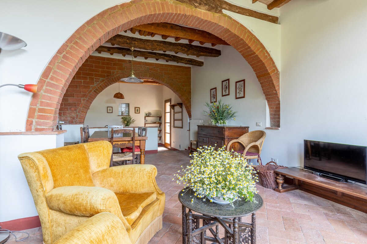 A spacious living area is highlighted by terracotta flooring and exposed wooden beams. Arched brick accents frame the doorway leading to the dining area, which features a wooden table and chairs. An antique sideboard and a cozy yellow armchair are positioned around a decorative table.