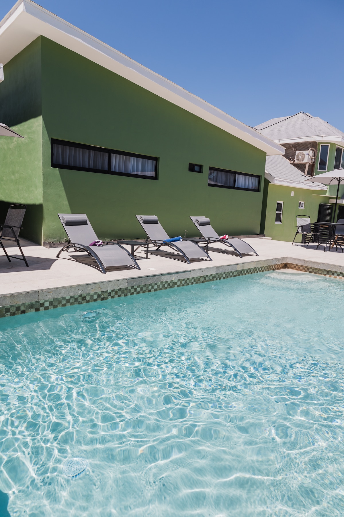 A clear swimming pool is framed by a light-colored deck area. Sun loungers are arranged along the edge, offering space for relaxation. The building's vibrant green exterior contrasts with the blue sky above, while large windows reflect the surrounding outdoor environment.