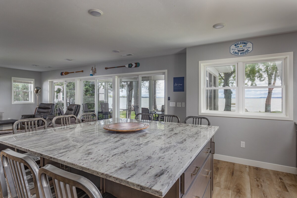 A spacious kitchen area features a large granite island with seating around it, providing an open view of the lake through multiple wall-length windows. Natural light enhances the neutral tones of the decor, with comfortable seating options visible in the background.