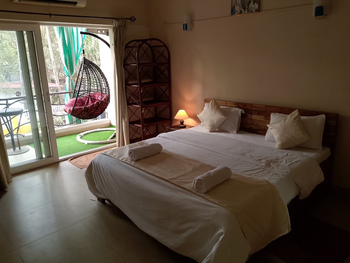 A spacious bedroom is equipped with a large bed dressed in crisp linen and two rolled towels. A cozy hanging chair is visible near a balcony with sliding glass doors, allowing natural light to fill the room. A soft lamp provides gentle illumination.