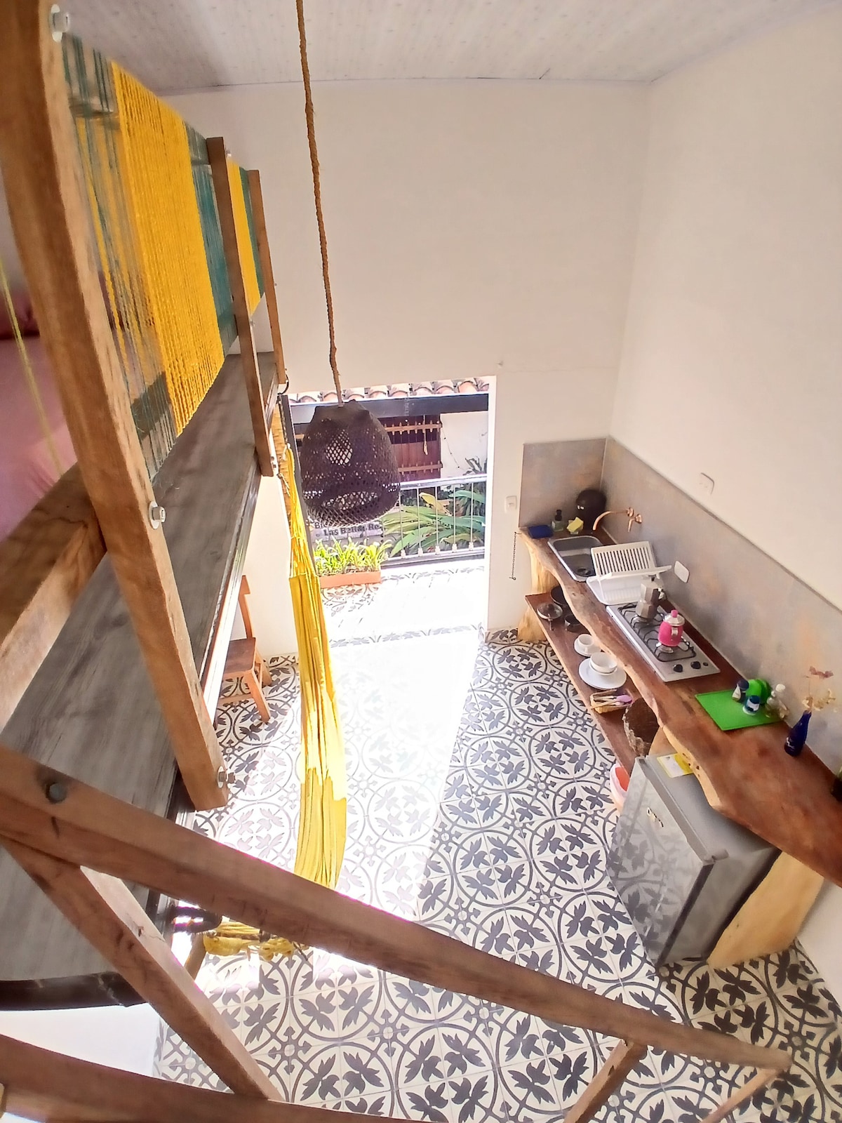 An overhead view of the kitchen area shows a blend of rustic wood and vibrant accents. Colorful tile flooring is highlighted, while a small dining space features a wooden table. Natural light enters through an open window, enhancing the inviting ambiance.