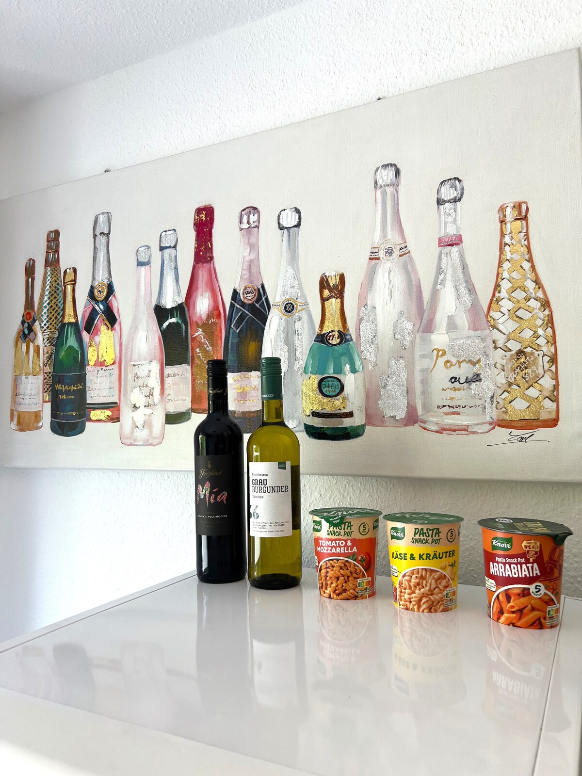 A collection of illustrated champagne bottles adorns the wall, providing a colorful backdrop. A selection of wine bottles in dark and light hues is placed on a sleek countertop, accompanied by various pasta dishes in packaged containers. The arrangement creates an inviting dining ambiance.