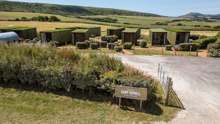 A view of Camp Tapnell showcases a cluster of glamping containers, nestled among greenery. The rolling countryside extends into the distance under a clear sky, inviting outdoor exploration and relaxation.