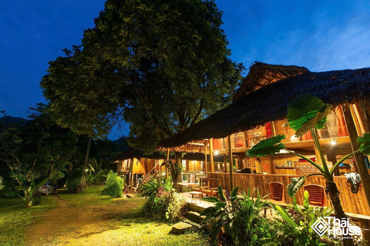 The exterior of Thai House is illuminated at night, showcasing its traditional wooden structure beneath a large tree. Lush greenery surrounds the area, enhancing the tranquil ambiance. The thatched roofs and wooden railings contribute to the rustic charm of this heritage accommodation.