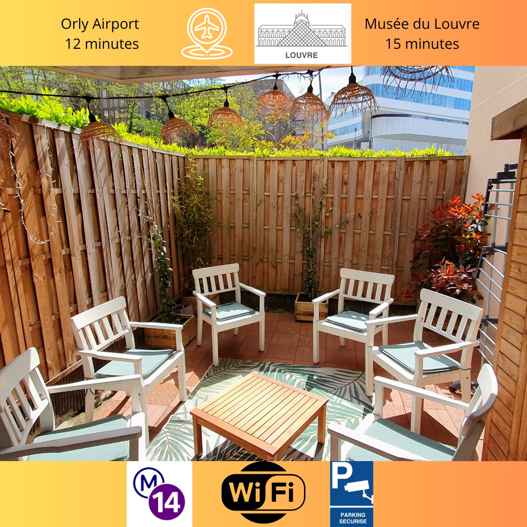 An outdoor terrace is featured, showcasing several white chairs arranged around a small wooden table. Lush greenery and decorative plants provide a natural ambiance, while wooden fencing ensures privacy. The area is well-lit, inviting relaxation and outdoor enjoyment.