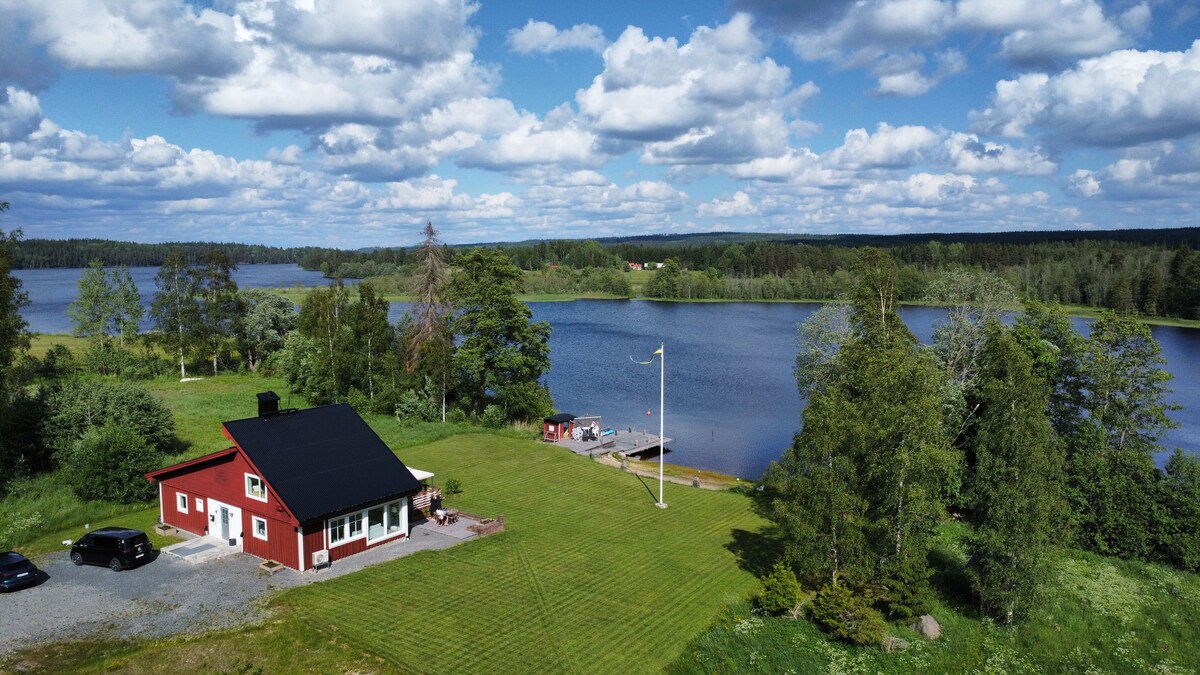 A newly renovated villa with a black roof is set on a spacious green lawn beside a serene lake. The property features a private dock and a picturesque view of the surrounding trees and water, providing a peaceful retreat in nature.