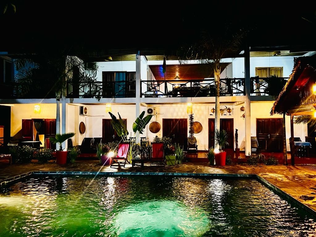 A well-lit villa features a spacious patio overlooking a pool. Chairs are positioned near the water, and tropical plants add a vibrant touch. The second floor boasts balcony access, inviting a sense of relaxation in this tranquil setting.