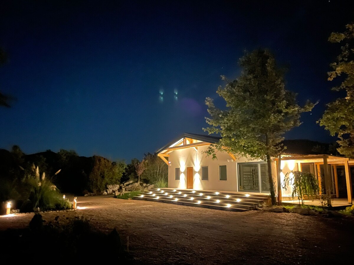 A well-lit home features a spacious entryway, complemented by soft lighting along the steps and pathway. Surrounding trees are enhanced by ground lights, creating a welcoming ambiance against the night sky.