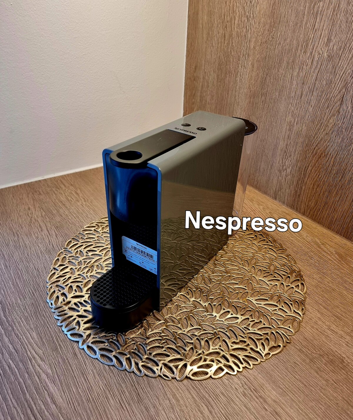 A modern Nespresso coffee machine is positioned on an intricately designed circular tray. The machine features a sleek metallic finish, with textured surfaces and a minimalist design, complementing the natural wood backdrop.