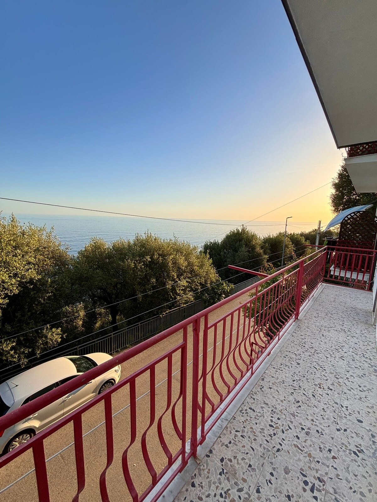 A balcony with a red railing offers views of the sea and surrounding greenery. The horizon is marked by a soft sunrise, casting gentle colors across the sky, while a street and parked car are visible below.