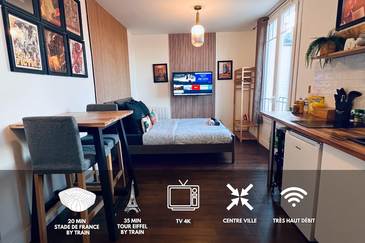 A modern studio space features a spacious bed covered with a textured blanket. A flat-screen 4K TV is mounted on the wall, while a small dining area with high chairs is positioned nearby. Natural light enters through large windows, enhancing the neutral color palette.