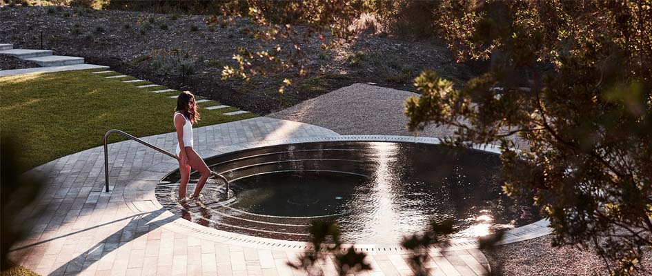 A serene outdoor spa is featured, with circular stones surrounding the water. A gentle staircase invites entry, while natural landscaped greenery and light create a tranquil atmosphere. Soft natural light reflects off the water's surface, enhancing the inviting setting.