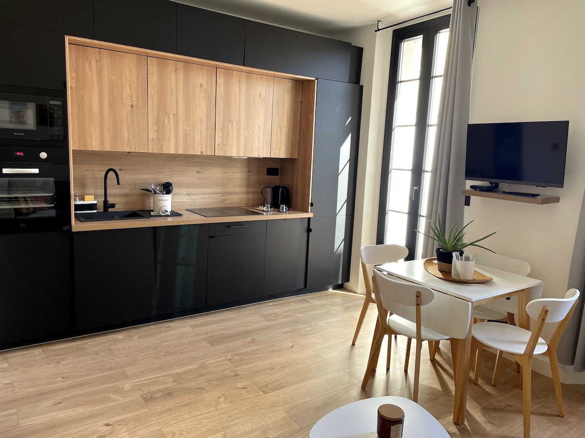 The kitchen features modern black cabinetry complemented by natural wood accents. A dining table with four chairs is positioned nearby, and a small television is mounted on the wall. Sunlight streams through the tall windows, illuminating the light wood flooring.