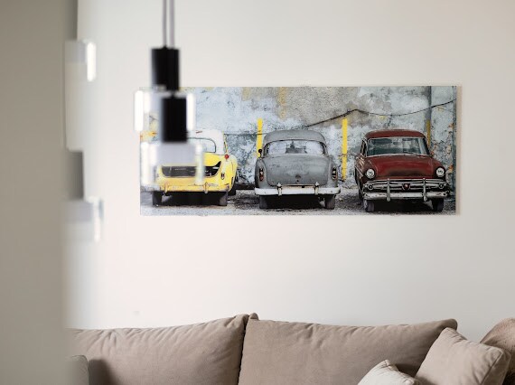 A wall-mounted artwork displays three vintage cars parked against a textured concrete backdrop. The yellow, gray, and red vehicles are positioned side by side, with bright yellow poles visible on either side, adding a playful contrast to the muted setting.