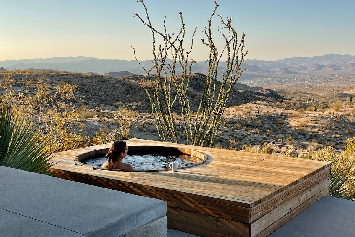 An outdoor cedar hot tub is situated on a wooden deck, offering a scenic view of the surrounding Mojave Desert terrain. Desert vegetation, including tall cacti, is visible nearby, with mountain ranges stretching into the distance under a clear sky.