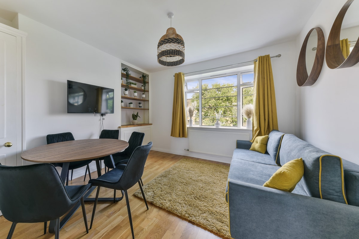 A living room features a comfortable blue sofa with accent cushions, a round wooden dining table surrounded by four black chairs, and a rug on a hardwood floor. Natural light floods the space through large windows, framed by yellow curtains.