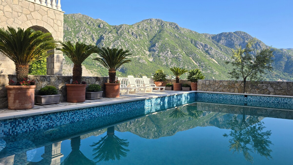 A serene swimming pool reflects the surrounding mountains, framed by lush palm trees in terracotta pots. Light-colored lounge chairs are arranged on the poolside, providing a peaceful space for relaxation. The vibrant blue tiles of the pool add a calm touch to the scenic backdrop.